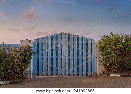 Curved Weathered Blue Wooden Garden Gate With Green Bushes At Both Sides And Partly Cloudy Sky At Su