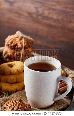 White Porcelain Cup Of Hot Tea, Cookies, Cinnamon Sticks Bunch On A Wooden Barrel Over Rustic Backgr