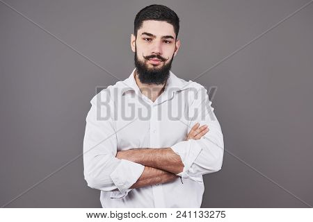Young Handsome Man Leaning Against Grey Wall With Arms Crossed. A Serious Young Man With A Beard Loo