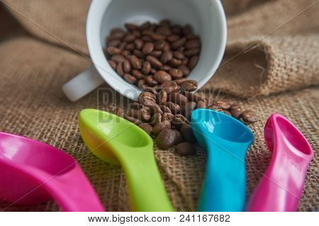 Loose Roasted Coffee Beans, White Porcelain Coffee Mug And Color Plastic Dose Measuring Spoons On Ju