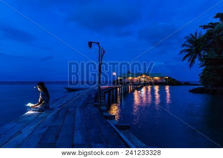 Woman sitting on a wooden pier and reading a book at dusk, using forehead light, Hatta island, Maluku, Indonesia