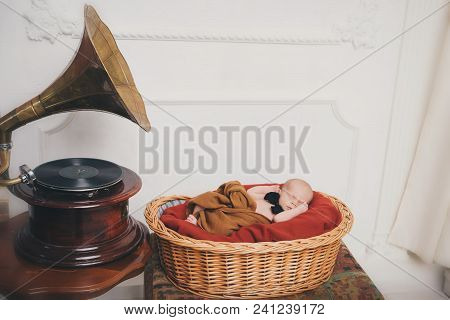 A Small Child With A Black Bowtie On His Neck In A Basket, Standing Next To The Phonograph