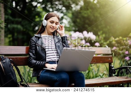 Smiling Student Sitting On Bench Listening Music And Holding Laptop In Park At University Preparing