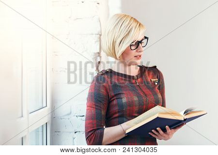 Pretty Intelligent Blonde Woman Wears Eyeglasses And Red Dress Reading Book Near Wide Window In Lagh