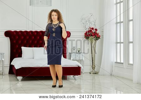 Woman in blue dress and black high-heel shoes stands in front of large red bed in bedroom.