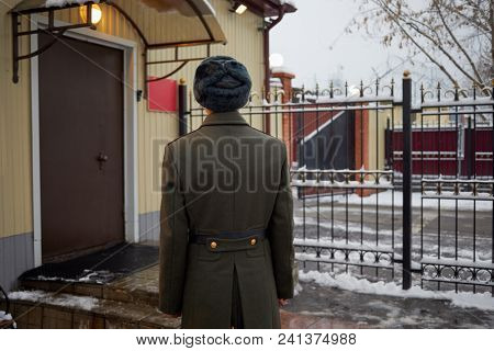 Young man in greatcoat stands near checkpoint office and metal fence on winter day, rear view.