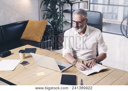Attractive And Confidental Adult Businessman Using Mobile Laptop Computer And Making Notes While Wor