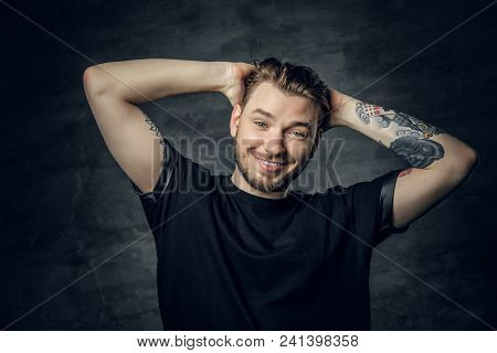 A Positive Hipster Male Dressed In A Black T Shirt Holds Head With Tattooed Arms.