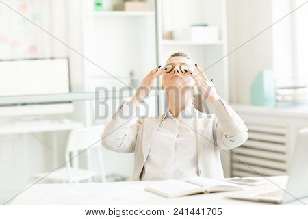 Young businesswoman with cucmber clices on her tired eyes relaxing by her workplace in office