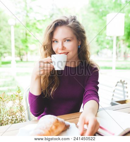 Beautifl Attractive Young Blonde Woman With Curly Hair Drinking Cappuccino And Eating Croissant In C