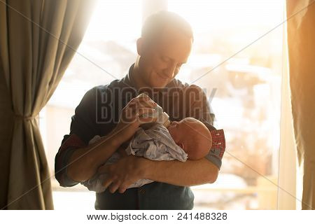 Happy Young Father Feeding Newborn Baby With Milk Bottle On Couch At Home