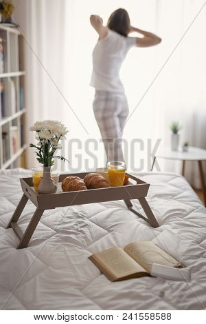 Breakfast Tray With Orange Juice, Croissants And Coffee Placed On Bed; Woman Standing Next To The Wi
