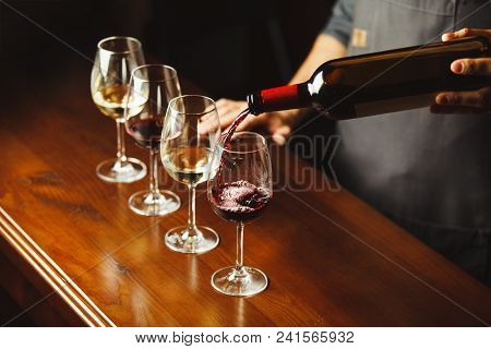 Bartender In Apron Pours Sweet Red Wine From Bottle In Capacious Glasses On Wooden Bar Counter. Deli