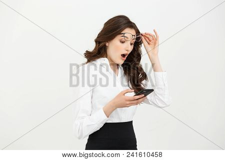 Business Concept: Surprised Youngwoman Holding Mobile Phone And Staring At It While Standing Against