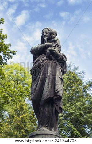 Old Statue Of Virgin Mary With Baby Jesus Christ Against Blue Sky. Stone Sculpture Of Mother Of God 
