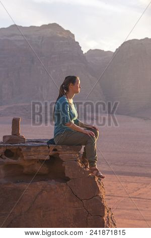 The Girl-traveler Sits On A Stone Ledge Against The Backdrop Of The Mountains In The Wadi Wadi Deser