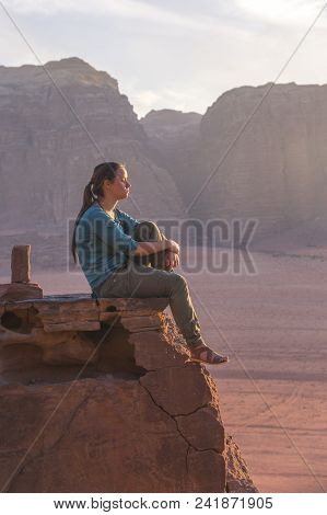The Girl-traveler Sits On A Stone Ledge Against The Backdrop Of The Mountains In The Wadi Wadi Deser