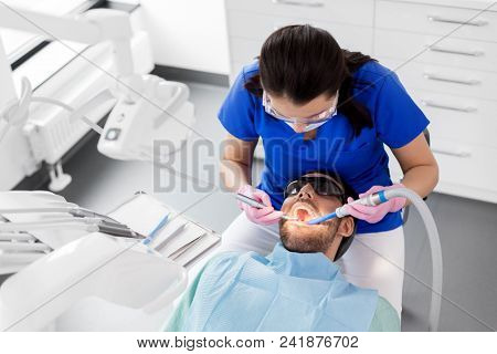 medicine, dentistry and healthcare concept - female dentist with drill and saliva ejector treating male patient teeth at dental clinic
