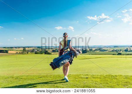 Full length rear view of an active woman carrying a blue stand bag, while walking on the grass towards a training area for professional golf in a sunny day