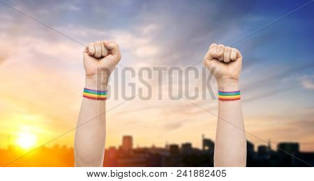 lgbt, same-sex relationships and homosexual concept - close up of male hands wearing gay pride awareness wristbands showing fist over city sunset background