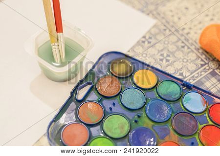 Two Paintbrushes Standing In A Plastic Container Of Water And A Box Of Multicolored Water Colour Pai