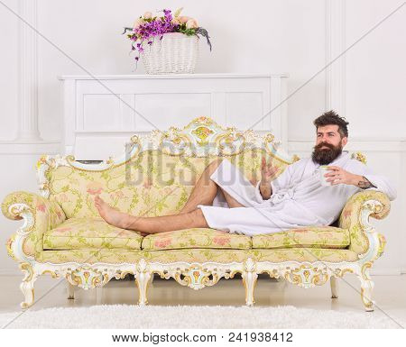 Man With Beard And Mustache Enjoys Morning While Lay On Luxury Sofa. Man On Sleepy Smiling Face In B