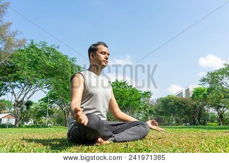 Indian Man Meditating In Lotus Pose Outdoors On Summer Lawn With Trees In Background. Outdoor Yoga C