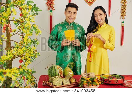 Cheerful Young Asian Couple In Room Decorated For Tet Celebration Standing At Table Served With Trad