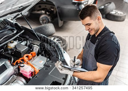 Smiling Mechanic Writing On Clipboard While Inspecting Car Engine Compartment