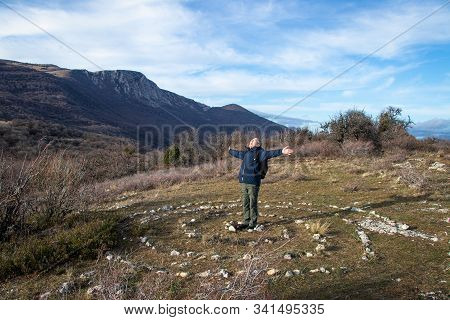 A Smiling Happy Adult Man, A Hiker, With Backpack, With His Arms Spread Wide, Stands In The Center O