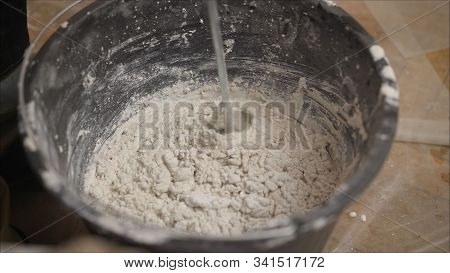 Builder Mixing Plaster In A Bucket Using An Electric Mixer. Builder Mixes The Mixture In A Bucket Us