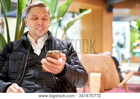 Sniling male in leather jacket with smartphone while sitting on chair in store against green plant