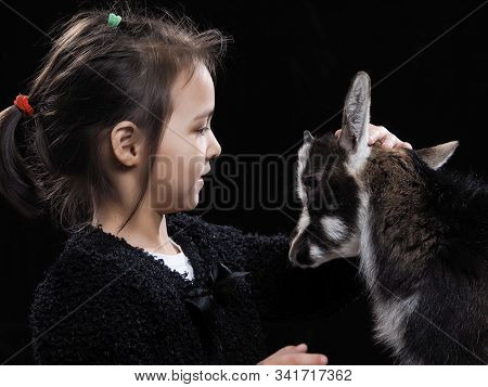A Girl Of Asian Appearance With A Small Goat On A Black Background. Portrait Of A Child With A Pet
