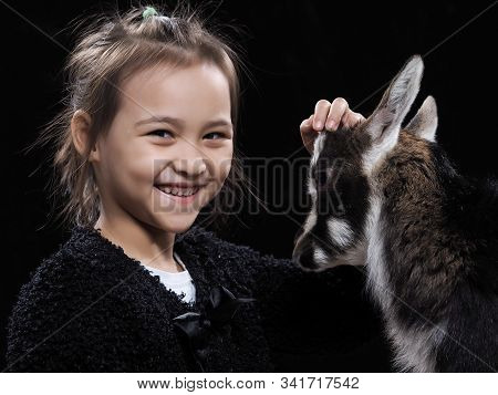 A Girl Of Asian Appearance With A Small Goat On A Black Background. Portrait Of A Child With A Pet