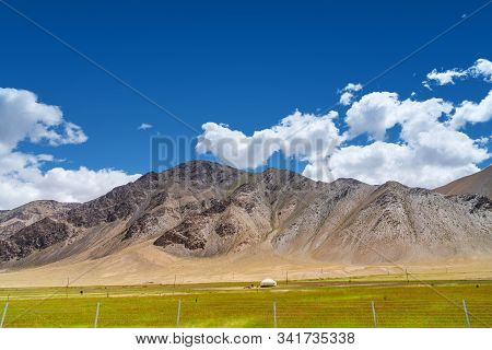 Vibrant Green Pasture With Yurt At The Foot Of Barren Rocky Mountains Under Cloudy Blue Sky In Pamir