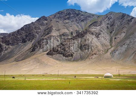 Vibrant Green Pasture With Yurt And Farmer Walking At The Foot Of Barren Rocky Mountains Under Cloud