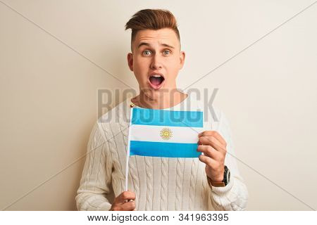 Young handsome man holding Argentinian Argentina flag over isolated white background scared in shock with a surprise face, afraid and excited with fear expression