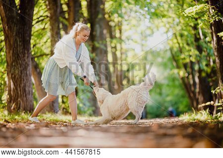 Happy Walk With Your Beloved Pet In Autumn Forest. Young Woman In Dress Is Playing With Labrodor Gol