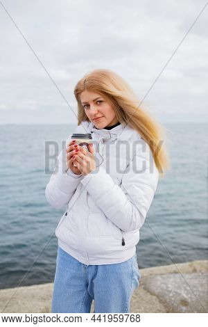 Young Blonde Lady Stands Near Seaside And Drink Coffee, It Is Cold Winter Ourside, Low Temperature. 