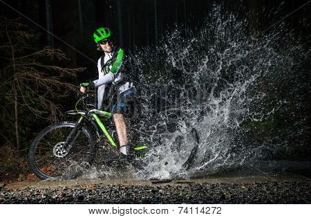 Mountain biker speeding through forest stream, water splash in freeze motion.