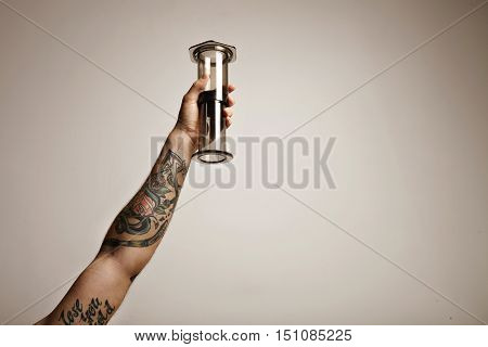 Close up shot of a tattooed man's hand holding clear light gray aeropress nontraditional coffee brewing equipment high in the air isolated on white