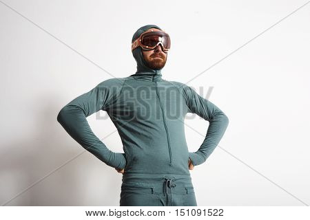 Young fitted bearded male athlete in baselayer thermal suite wears snowboarding googles, proudly stands in front of white wall