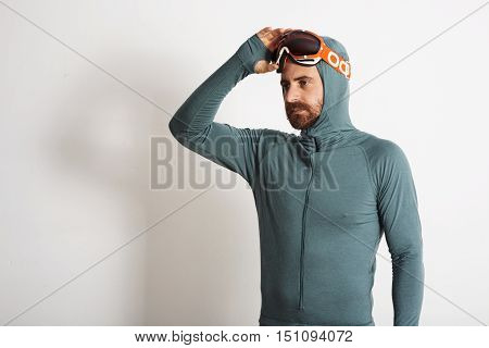 Young fitted bearded male athlete in baselayer thermal suite removes his snowboarding googles with one hand, isolated on white background