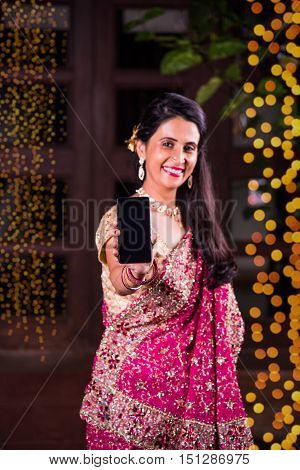 Young indian smart girl or lady showing smart phone screen against diwali lighting  background. indian woman celebrating diwali with smartphone showing blank screen