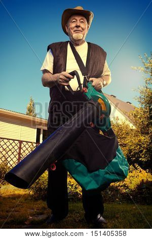 Autumn in a garden. Senior man removes fallen leaves in his garden using leaf blower.