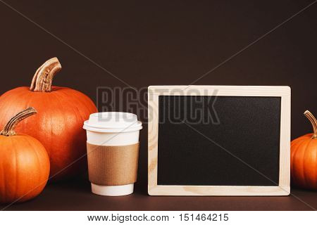 Coffee to take away with spices and pumpkin. Concept of traditional autumn spicy latte. Place for logo on brown coffee holder. Black chalk desk with wooden frame is for placing your text.