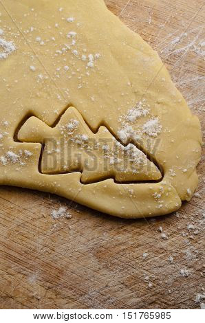 Christmas tree shape cut out from biscuit or cookie dough on old wooden chopping board with scattered flour sprinkles.