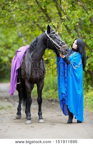 Black-haired woman in blue capote stands with horse in the park.