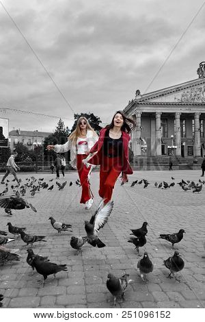 Two Fashion Young Girl Running And Lauging Acros The Square. Stylish Woman In A Red Suit And Sunglas