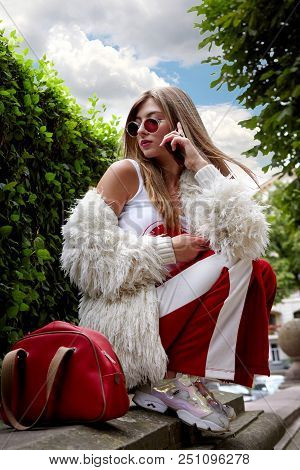 Fashion Young Girl Siting On The Border. Stylish Woman In White Fur And Sport Red Pants With Red Woo